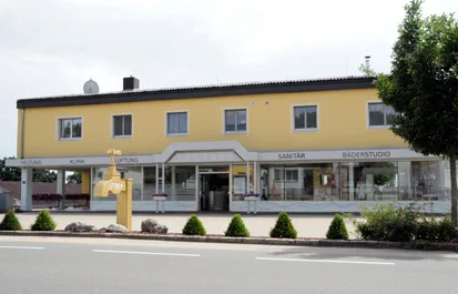 Das Lettner-Gebäude in gelber Fassung mit Klima, Lüftung, Sanitär und Bäderstudio, davor der goldene Wasserhahn