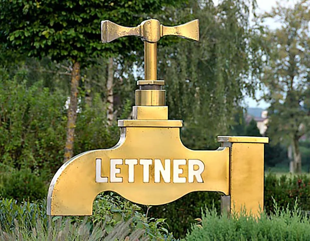 Goldener Riesen-Wasserhahn mit dem Schriftzug LETTNER vor Bäumen
